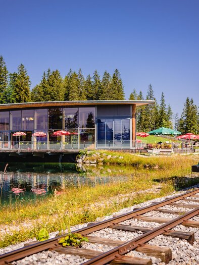 A green railway by a pond, next to a modern building with large windows. In the background, there are trees and some tables with umbrellas. | © Mariazeller Bürgeralpe / Rudy Dellinger