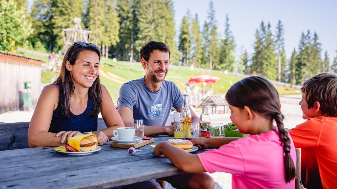 Eine Familie genießt ein Frühstück im Freien. Im Hintergrund sind Bäume und eine sonnige Landschaft zu sehen. | © Mariazeller Bürgeralpe / Rudy Delllinger
