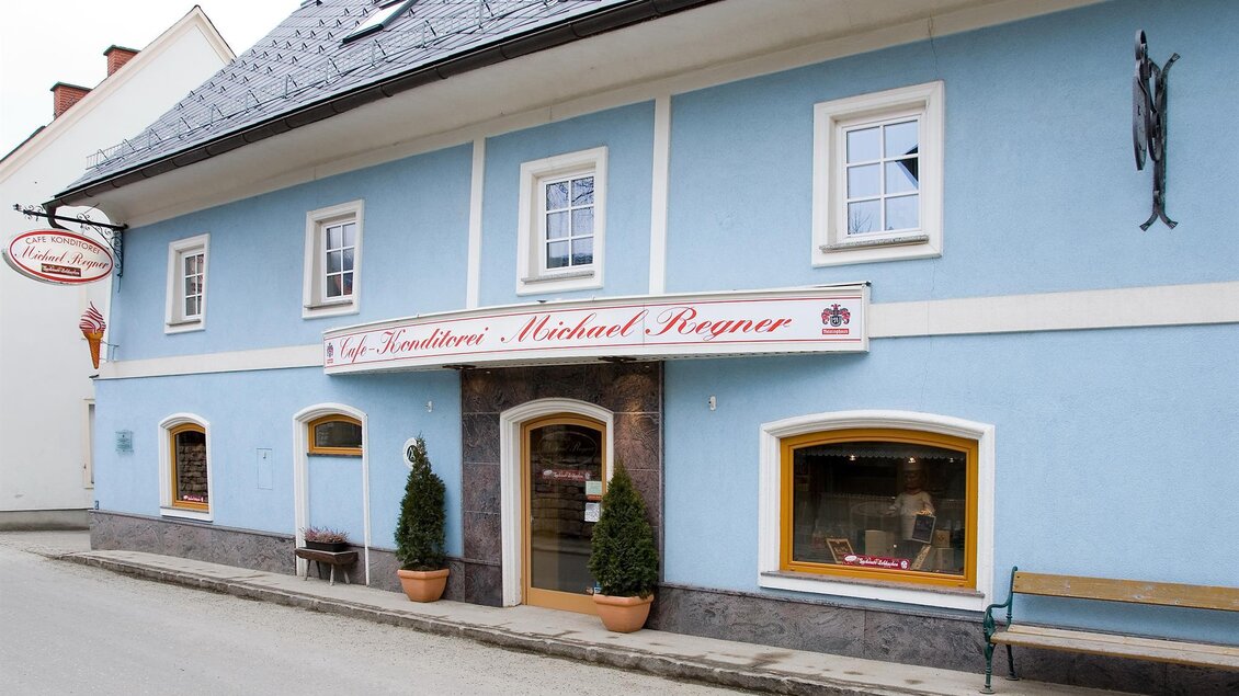 Ein blaues Gebäude mit einem gemütlichen Eingang und einem Schild, das einen Namen trägt. Vor dem Gebäude stehen kleine Pflanzen und eine Bank. | © Konditorei Regner