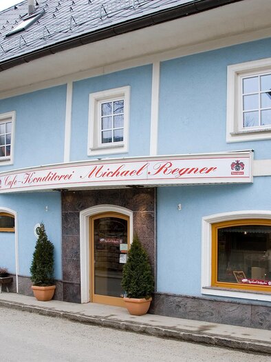 Ein blaues Gebäude mit einem gemütlichen Eingang und einem Schild, das einen Namen trägt. Vor dem Gebäude stehen kleine Pflanzen und eine Bank. | © Konditorei Regner
