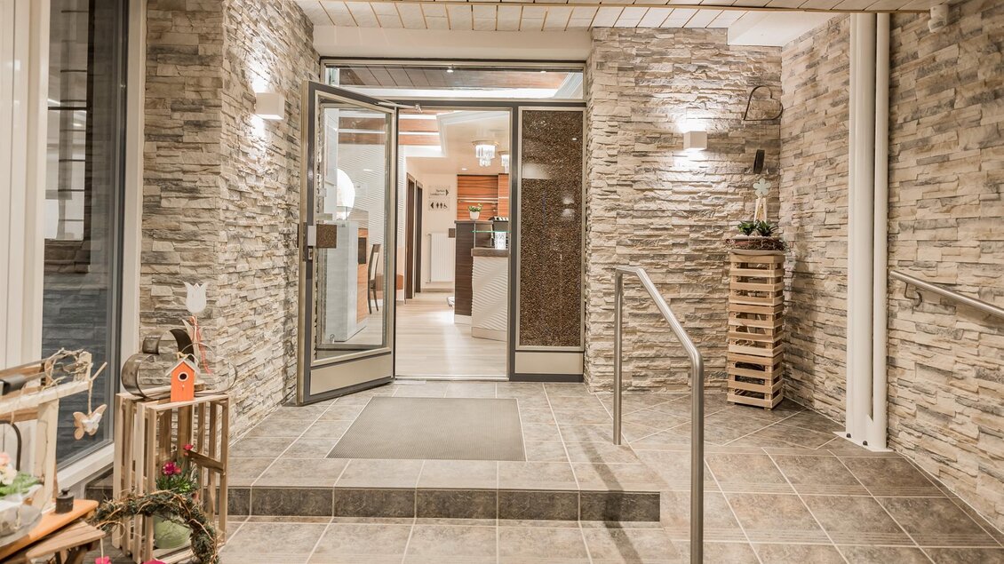 Ein einladender Eingangsbereich mit modernen Steinwänden und einer gläsernen Tür. Der Boden ist mit großen Fliesen gestaltet und in der Nähe stehen Dekorationselemente. | © Konditorei Regner