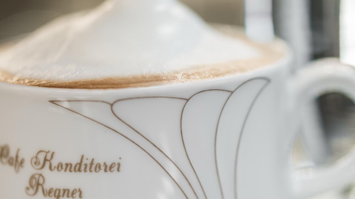 Eine Tasse Kaffee mit feinem Schaum und einem Zuckergefäß daneben. Im Hintergrund ist ein Glas Wasser zu sehen. | © Konditorei Regner