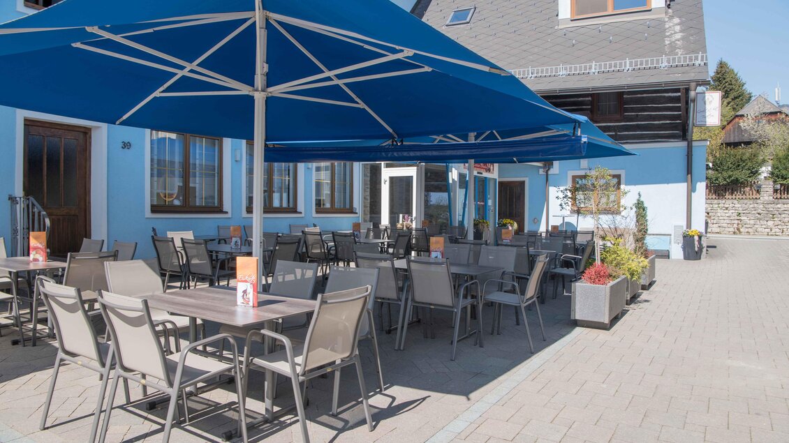Ein gemütlicher Außenbereich mit Tischen und Stühlen unter einem blauen Sonnenschirm. Die Umgebung ist hell und einladend. | © Konditorei Regner