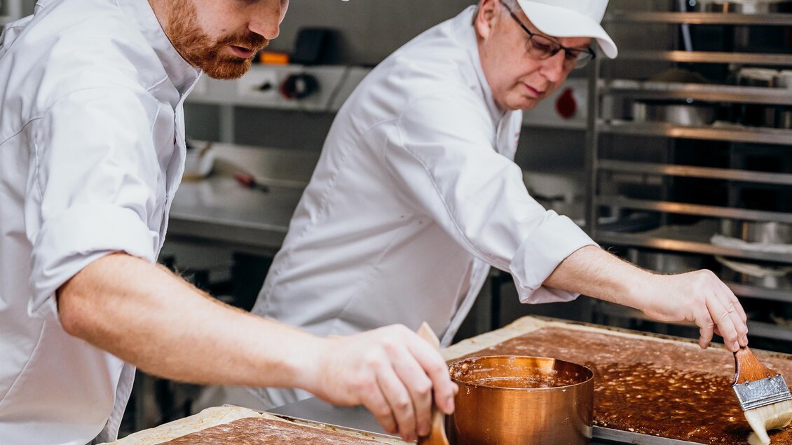 Zwei Bäcker arbeiten in einer modernen Backstube. Sie bereiten Schokoladenteig auf großen Tabletts vor. | © Konditorei Regner