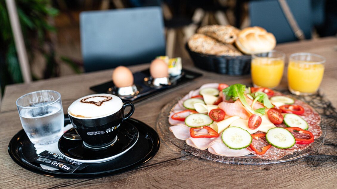 Ein Frühstückstisch mit einem Cappuccino, zwei Eiern, frischem Gemüse und einem Obstsalat. Im Hintergrund sind Brötchen und ein Glas Orangensaft zu sehen. | © Kalthuber photography