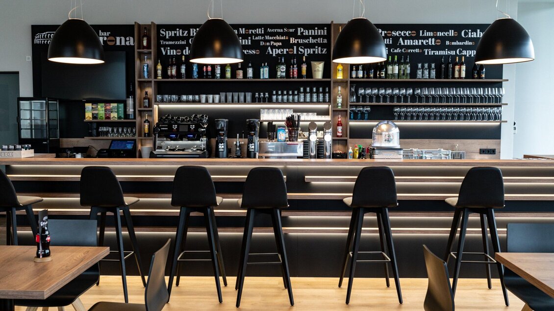 Eine moderne Bar mit einem langen Holztresen und hohen Stühlen. Im Hintergrund sind Regale mit verschiedenen Getränken und einer gemütlichen Beleuchtung. | © Kalthuber photography