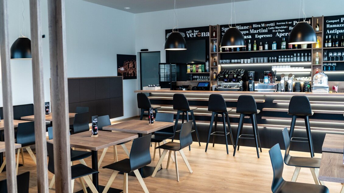 Ein modernes Restaurant mit Holztischen und schwarzen Stühlen. Die Bar im Hintergrund ist gut beleuchtet und bietet eine Auswahl an Getränken. | © Kalthuber photography