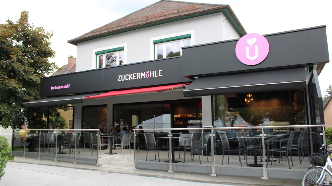 Das Bild zeigt ein Restaurant namens "Zuckermühle" mit einer modernen Fassade. Vor dem Gebäude gibt es eine Terrasse mit Sitzgelegenheiten und einen Blick auf die Straße. | © Rene Lederer