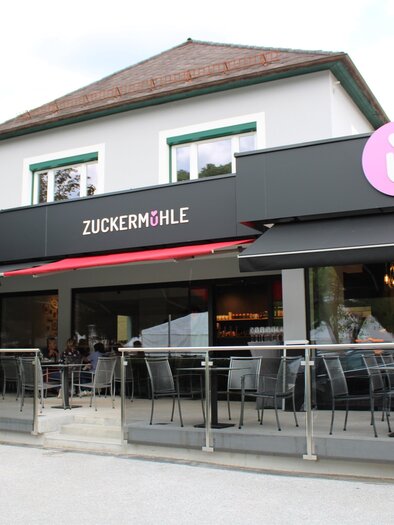 Das Bild zeigt ein Restaurant namens "Zuckermühle" mit einer modernen Fassade. Vor dem Gebäude gibt es eine Terrasse mit Sitzgelegenheiten und einen Blick auf die Straße. | © Rene Lederer