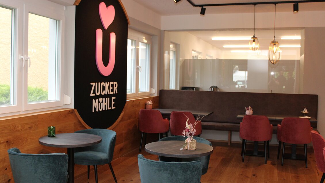 Ein einladendes Café mit gemütlichen Tischen und bunten Stühlen. An der Wand befindet sich ein auffälliges Schild mit der Aufschrift "Zucker Mühle". | © Rene Lederer