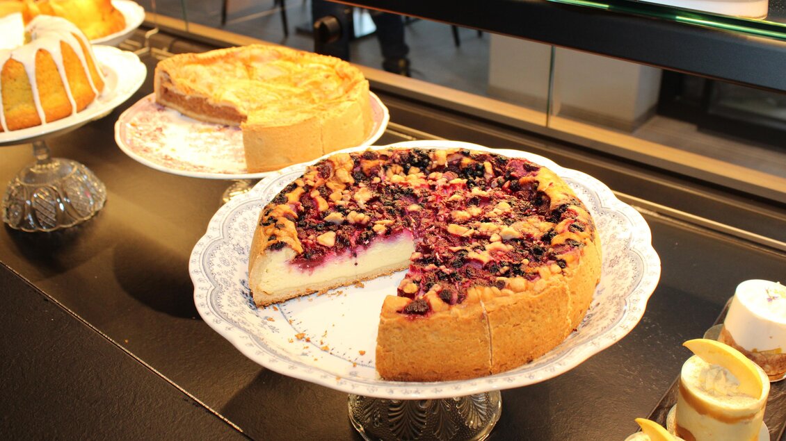 Ein köstlicher Kuchen mit einer fruchtigen Füllung und knackigem Belag steht auf einem dekorativen Teller. Im Hintergrund sind weitere Kuchen und Gebäck sichtbar. | © Rene Lederer