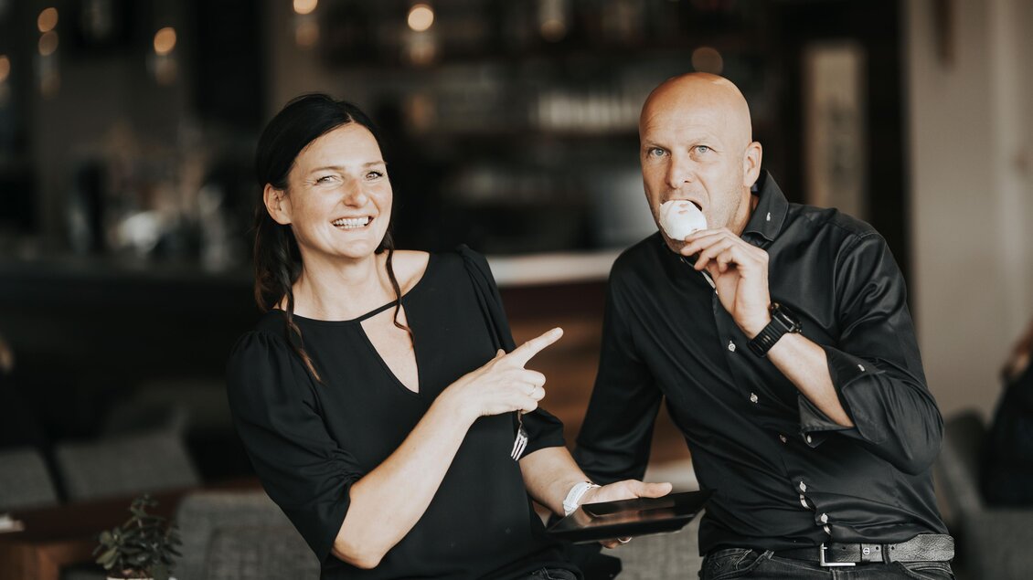 Ein Paar lacht gemeinsam in der Zuckermühle Bärnbach. Die Frau zeigt auf den Mann, der ein Eis isst. Beide tragen schwarze Kleidung in einem modernen Café-Ambiente. | © Die Abbilderei
