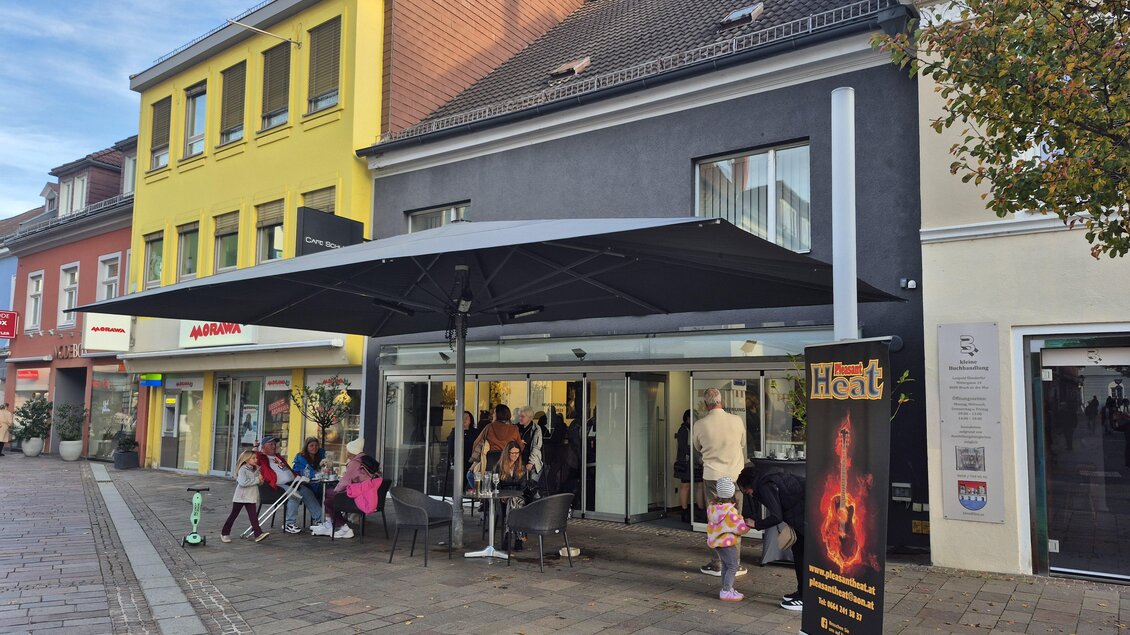 Ein modernes Café mit großzügiger Terrasse und Sonnenschirm. Menschen sitzen entspannt auf den Stufen und genießen die Atmosphäre in der Stadt. | © TV Hochsteiermark