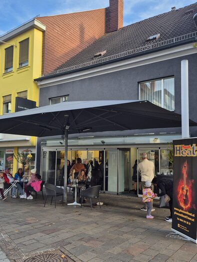 Ein modernes Café mit großzügiger Terrasse und Sonnenschirm. Menschen sitzen entspannt auf den Stufen und genießen die Atmosphäre in der Stadt. | © TV Hochsteiermark
