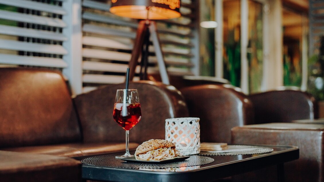 Ein eleganter Tisch mit einem Getränk und einem Snack. Im Hintergrund sind gemütliche Sofas und eine stilvolle Lampe zu sehen. | © GenussReich
