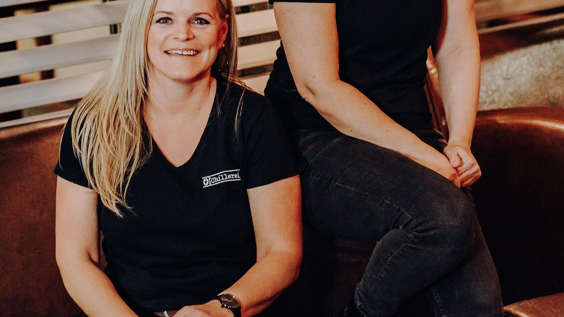 Zwei Frauen sitzen lächelnd auf einer Couch. Sie tragen schwarze T-Shirts und befinden sich in einem modernen, freundlichen Ambiente. | © GenussReich