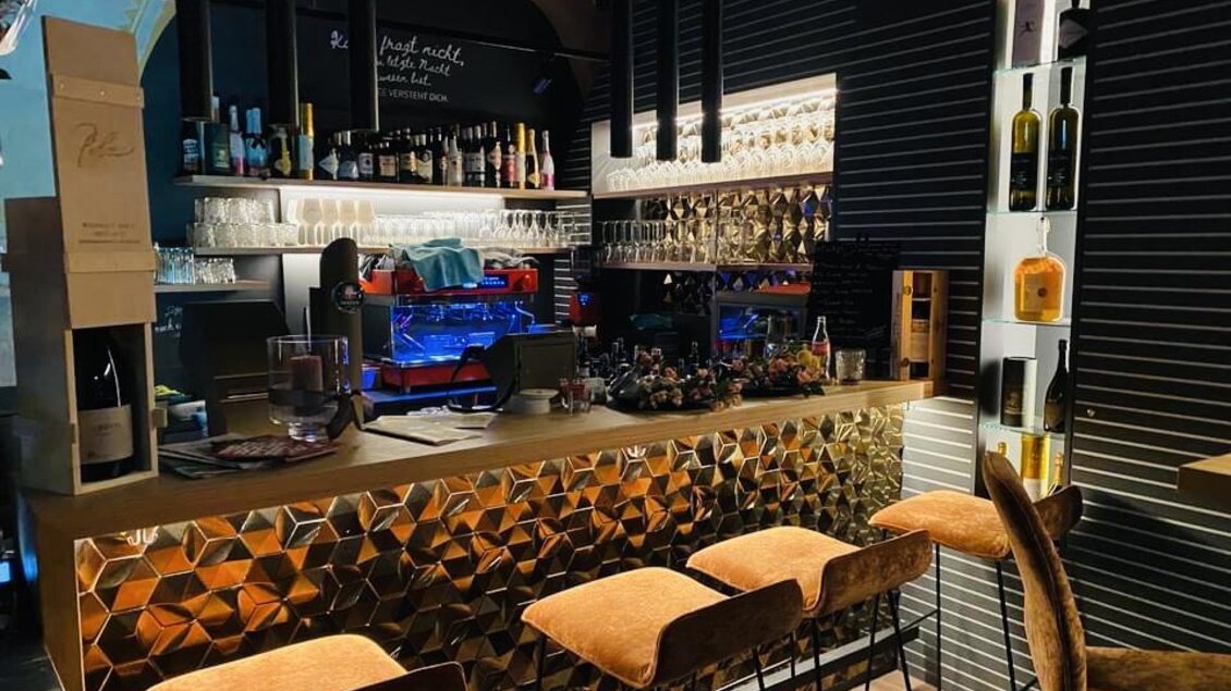 Eine stilvolle Bar mit einer goldenen Theke und gemütlichen Sitzplätzen. Im Hintergrund sind Regale mit einer Auswahl an Getränken und Beleuchtung zu sehen. | © Amaro