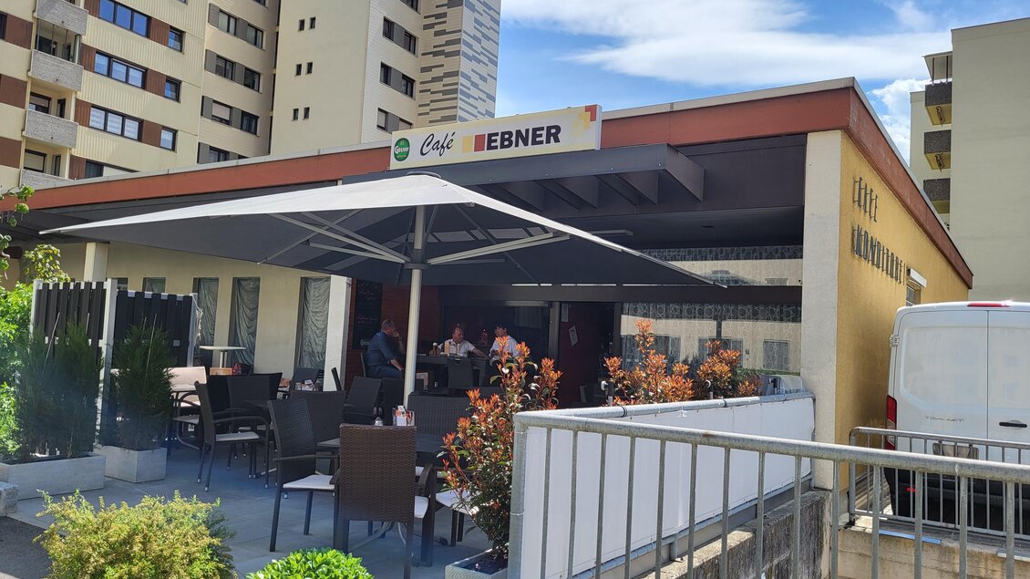 Ein Café mit einem überdachten Außenbereich und Sitzplätzen. Im Hintergrund sind mehrere Wohngebäude sichtbar. | © TV Hochsteiermark