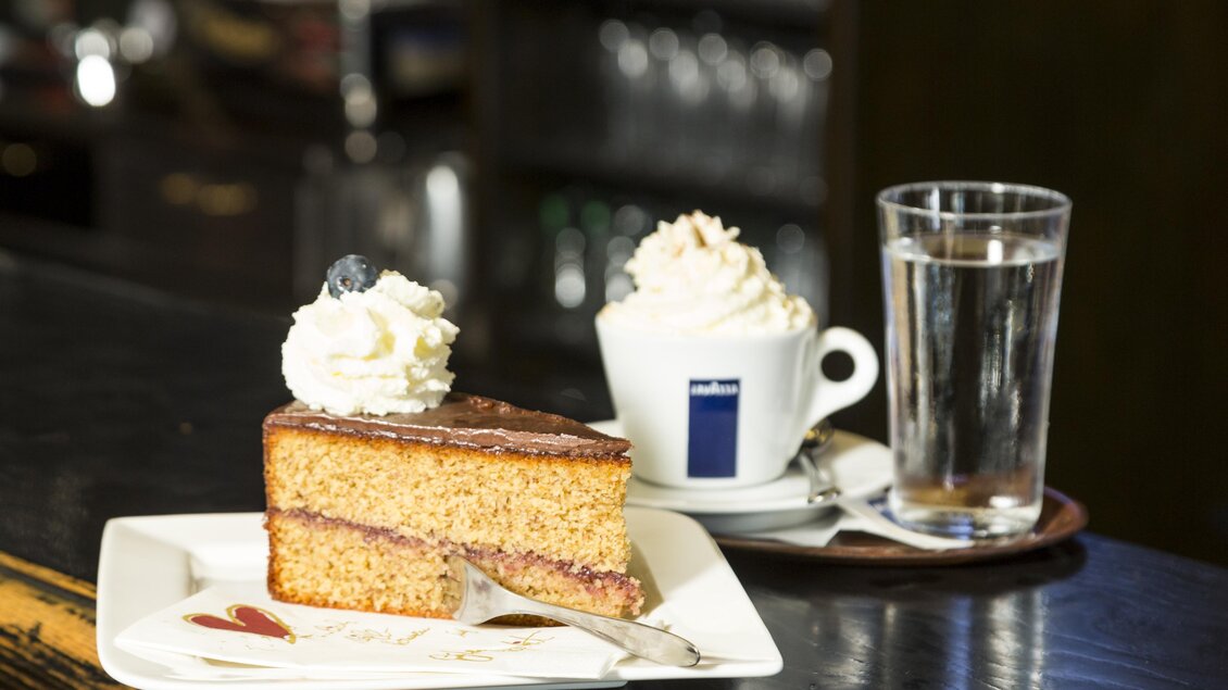 Ein Stück Kuchen mit Sahne und ein Becher Kaffee neben einem Glas Wasser. Die Anordnung ist auf einem Holztisch bei einem Café zu sehen. | © TV Region Graz - Rene Vidalli