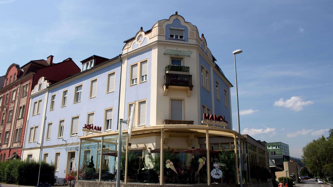 Ein historisches Gebäude mit blauer Fassade und einem Erdgeschoss mit großen Fenstern. Es befindet sich an einer Straßenecke und hat einen Balkon im oberen Stockwerk. | © C. Stein