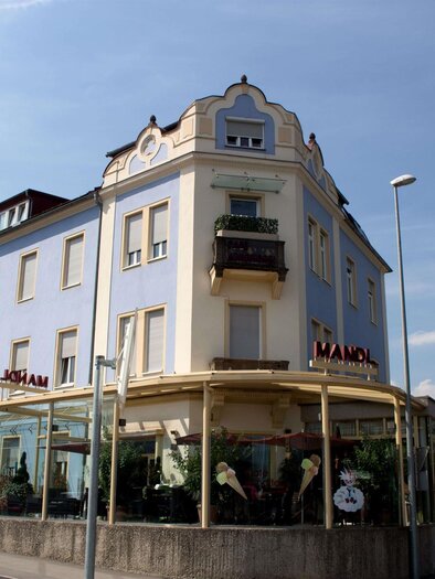 Ein historisches Gebäude mit blauer Fassade und einem Erdgeschoss mit großen Fenstern. Es befindet sich an einer Straßenecke und hat einen Balkon im oberen Stockwerk. | © C. Stein