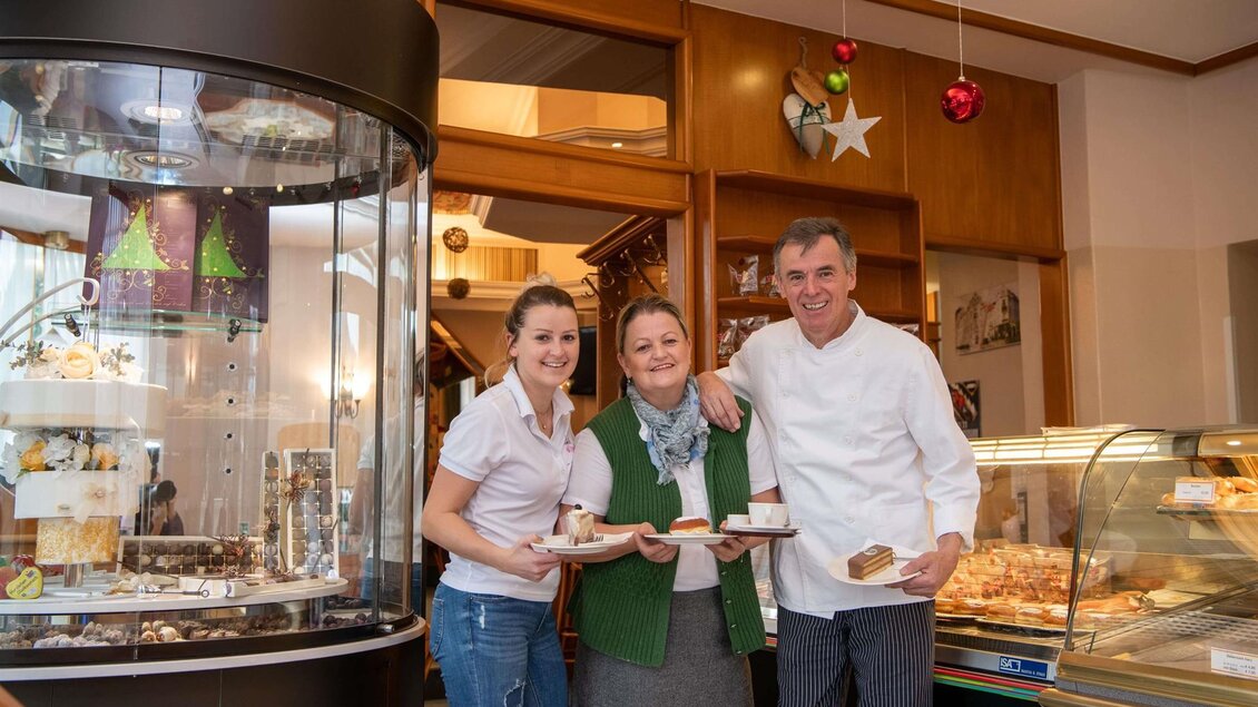 Ein freundliches Team von drei Personen steht in einer Bäckerei. Im Hintergrund sind köstliche Backwaren und festliche Dekoration zu sehen. | © K. Pashkovskaya