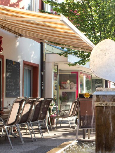 A cozy street café with several tables and chairs. In the foreground stands an elegant sculpture, while some guests relax. | © C. Stein