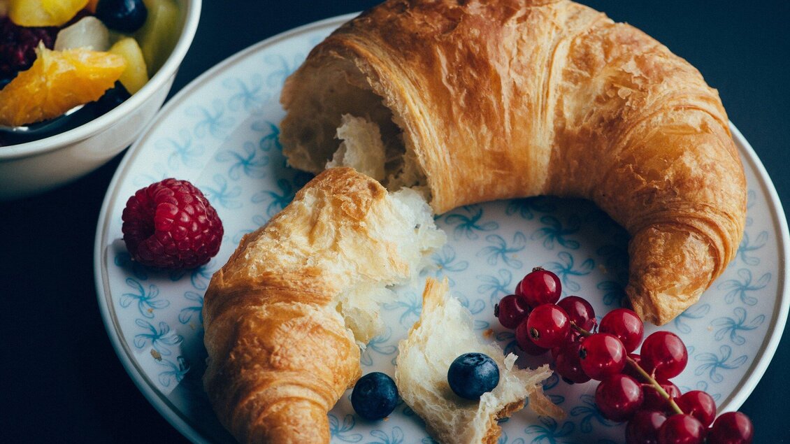 Ein frischer Croissant auf einem Teller, umgeben von Beeren und einer Schüssel mit frischem Obst. Der Croissant ist teilweise zerbrochen und zeigt die fluffige Innenstruktur. | © Cafe Promenade - Joel Kernasenko