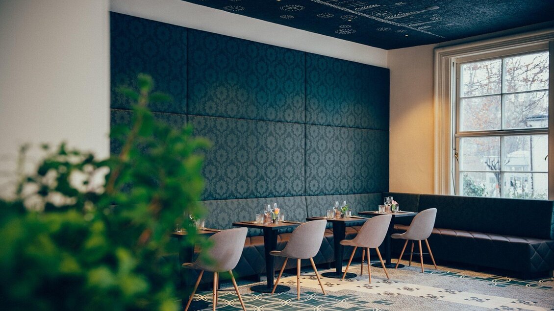 Ein modernes Restaurant mit eleganten Tischen und Stühlen. Die Wände sind in einem dunklen Muster gehalten und das Fenster lässt viel natürliches Licht herein. | © Cafe Promenade - Joel Kernasenko