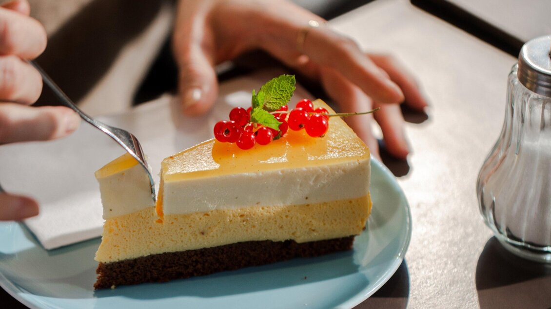 Ein Stück Käsekuchen auf einem blauen Teller, dekoriert mit roten Johannisbeeren und einem Minzblatt. Eine Hand hält einen Löffel, bereit, ein Stück des Kuchens zu nehmen. | © Cafe Promenade - Bobbys Agency