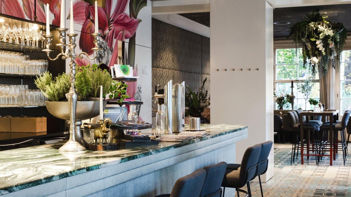 Eine elegante Bar mit einer marmorierten Theke und vielen Gläsern. Im Hintergrund sind Tische und ein schöner Pflanzendekoration zu sehen. | © Cafe Promenade - Bobbys Agency