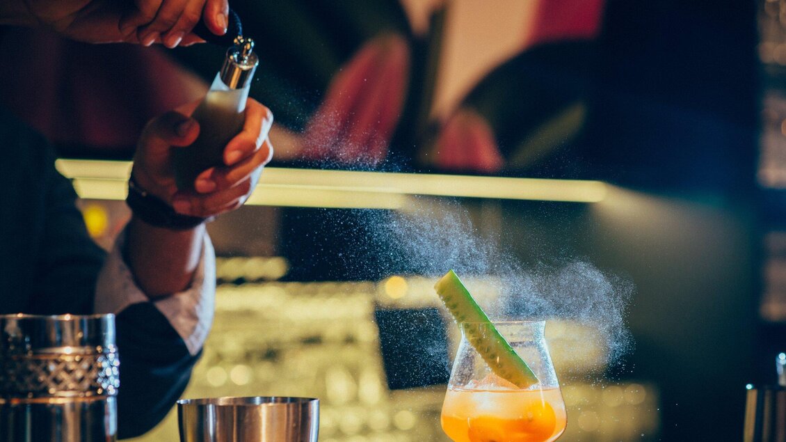 Ein Barkeeper bereitet einen Cocktail mit einer grünen Garnitur zu. Die Atmosphäre ist lebhaft und die Bar ist gut beleuchtet. | © Cafe Promenade - Joel Kernasenko