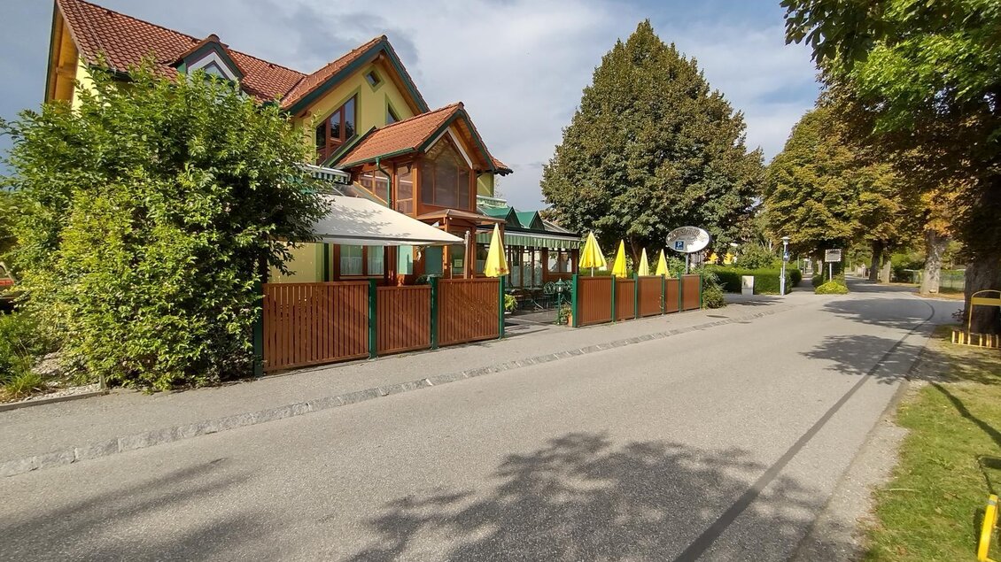 Ein gemütliches Haus mit einem Restaurantbereich und gelben Sonnenschirmen. Die ruhige Straße ist von Bäumen gesäumt. | © Thermen- und Vulkanland