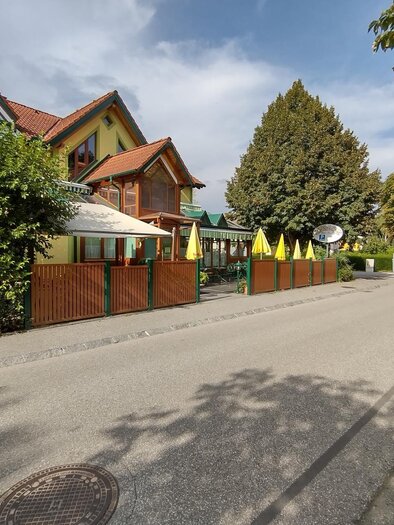 Ein gemütliches Haus mit einem Restaurantbereich und gelben Sonnenschirmen. Die ruhige Straße ist von Bäumen gesäumt. | © Thermen- und Vulkanland