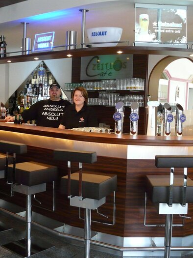 Eine moderne Bar mit Holztheke und Hockern. Zwei Mitarbeiter stehen lächelnd hinter dem Tresen. | © Cafe Zeitlos