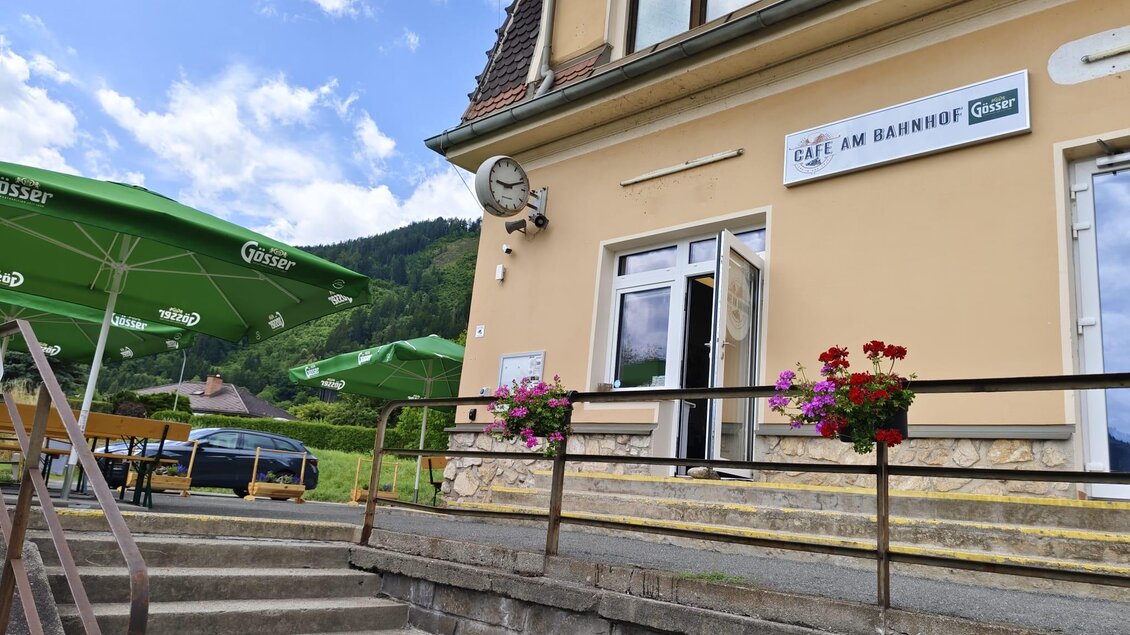 Ein gemütliches Café mit dem Namen "Café am Bahnhof" und blühenden Blumen. Im Hintergrund sind grüne Hügel und ein blauer Himmel zu sehen. | © SV Hinterberg