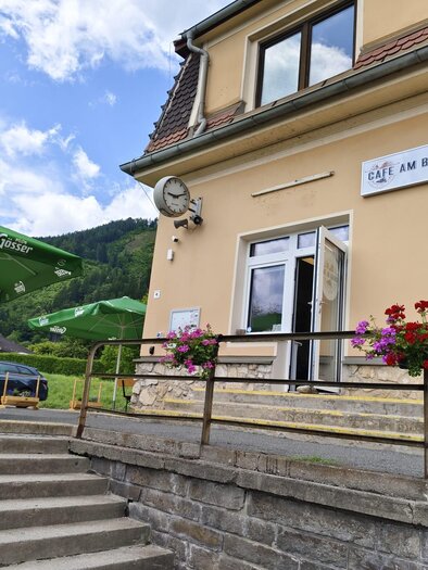 A cozy café named "Café am Bahnhof" with blooming flowers. In the background, green hills and a blue sky can be seen. | © SV Hinterberg