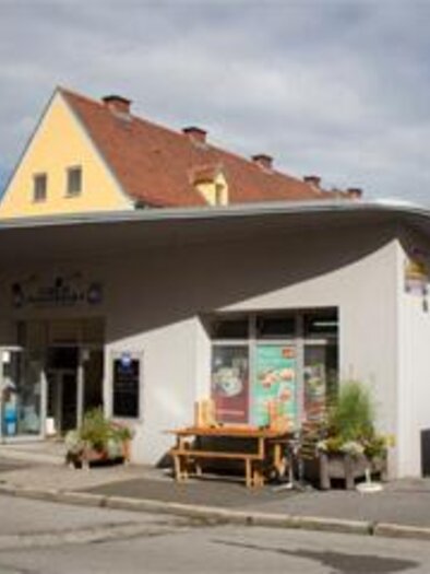 A small shop on a street corner with large windows.  
Outside there are plants and there are bicycles in front of the door. | © C. Stein