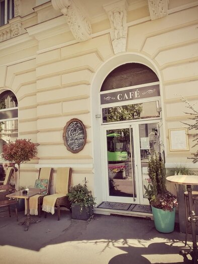 A cozy café with an inviting outdoor seating area. There are plants and an advertisement for ice cream next to the entrance. | © Julia Schoster