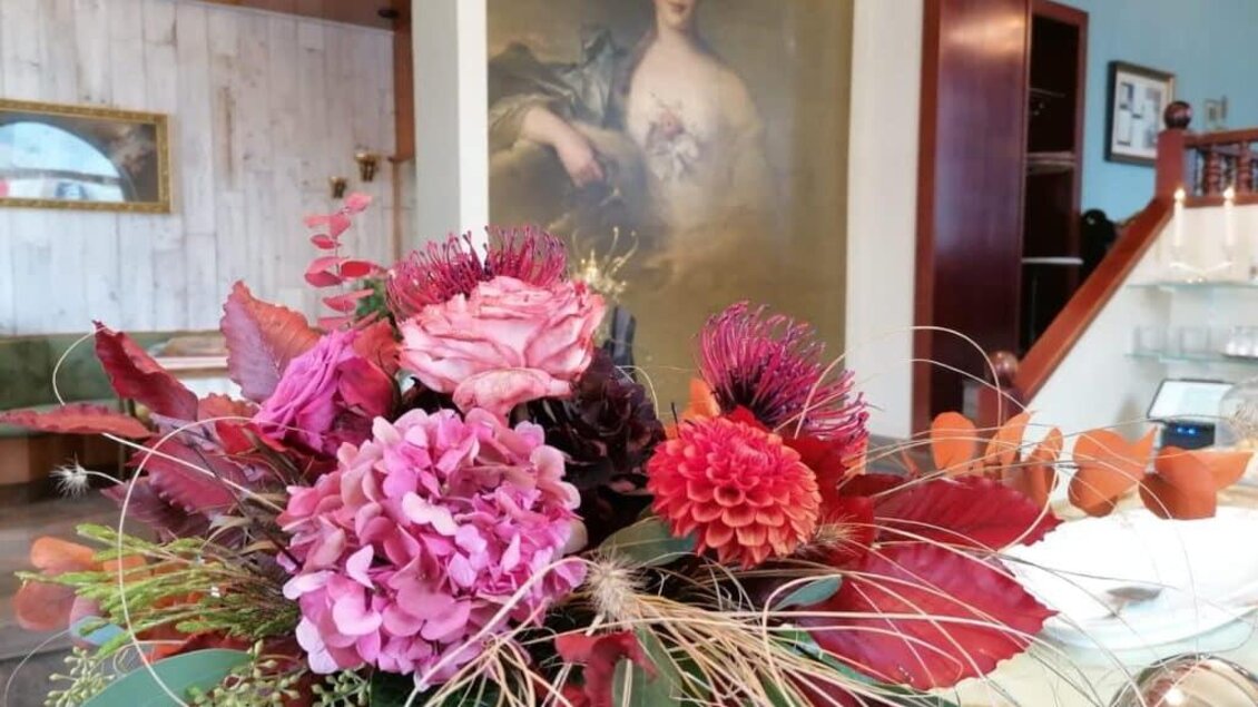 Ein bunter Blumenstrauß mit rosa und roten Blüten steht im Vordergrund. Im Hintergrund ist ein beeindruckendes Porträt einer Frau zu sehen. | © Anton Brandner