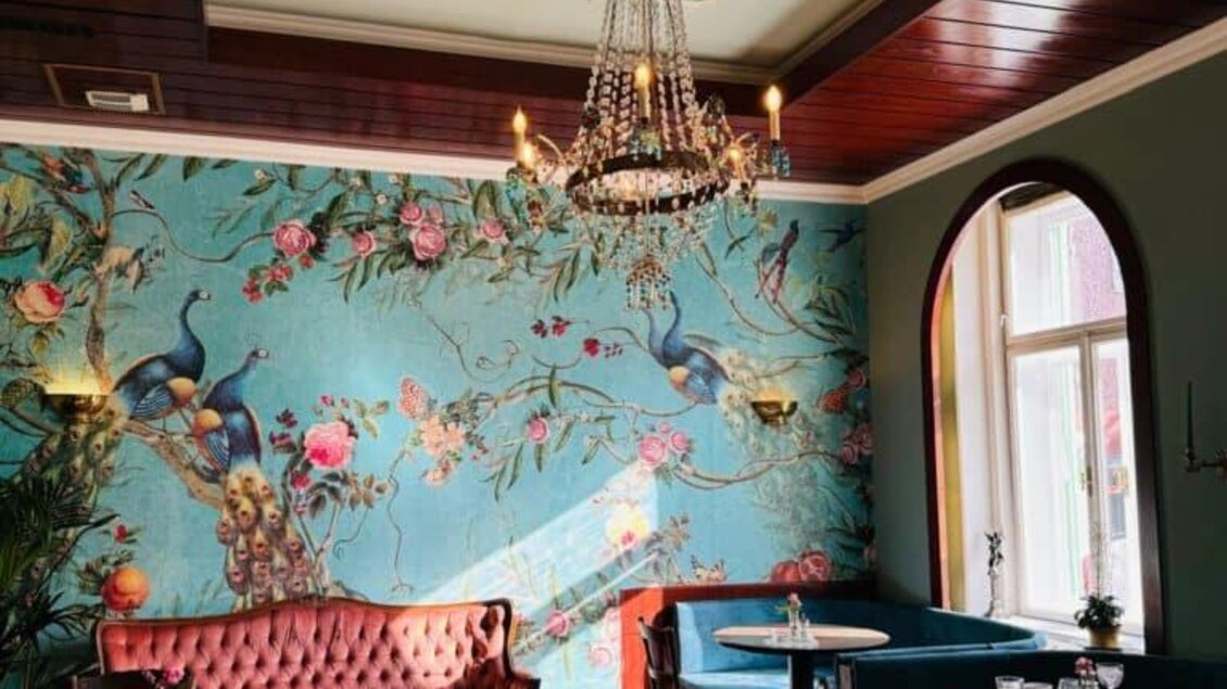 Ein stilvolles Café mit einer blauen Wandtapete, die Blumen und Vögel zeigt. Gemütliche Sitzgelegenheiten und ein eleganter Kronleuchter ergänzen das Ambiente. | © Anton Brandner