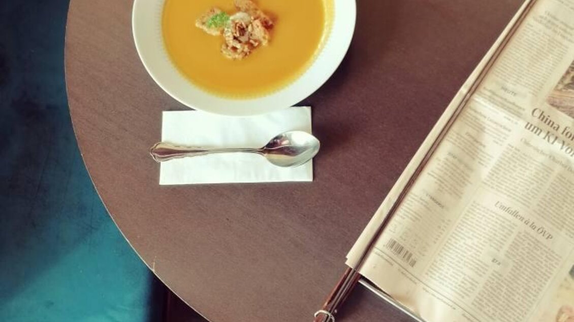 Eine Schüssel mit Kürbissuppe steht auf einem Holztisch. Daneben liegt eine Zeitung und ein Löffel auf einer Serviette. | © Anton Brandner