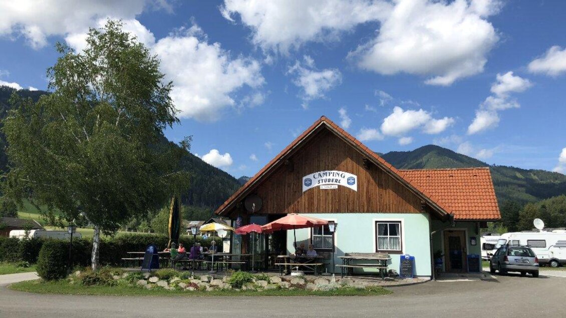 Ein großes Gebäude mit einem roten Dach und einer Terrasse. Umgeben von grünen Bäumen und Bergen unter einem blauen Himmel.