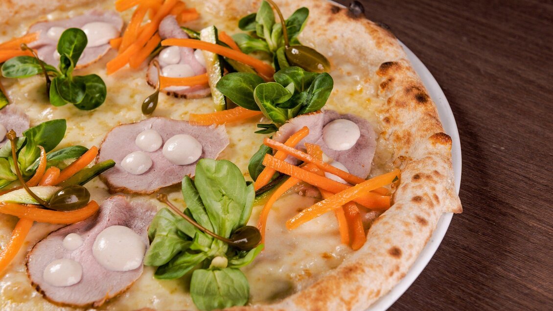 Eine leckere Pizza mit Schinken, frischen Kräutern und buntem Gemüse. Die Zutaten sind ansprechend angeordnet und verleihen der Pizza einen frischen Look. | © Casa Costiera