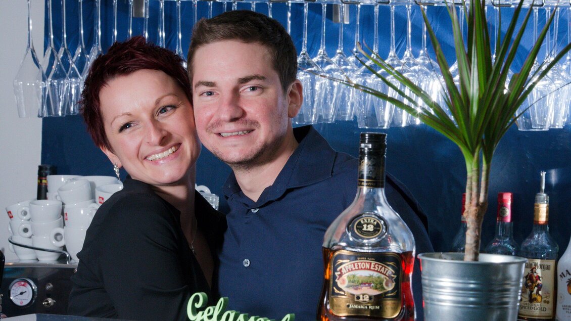 Ein fröhliches Paar steht hinter einer Bar, umgeben von Gläsern und Spirituosen. Sie lächeln freundlich in die Kamera. | © Caylend - Michael Fiedler