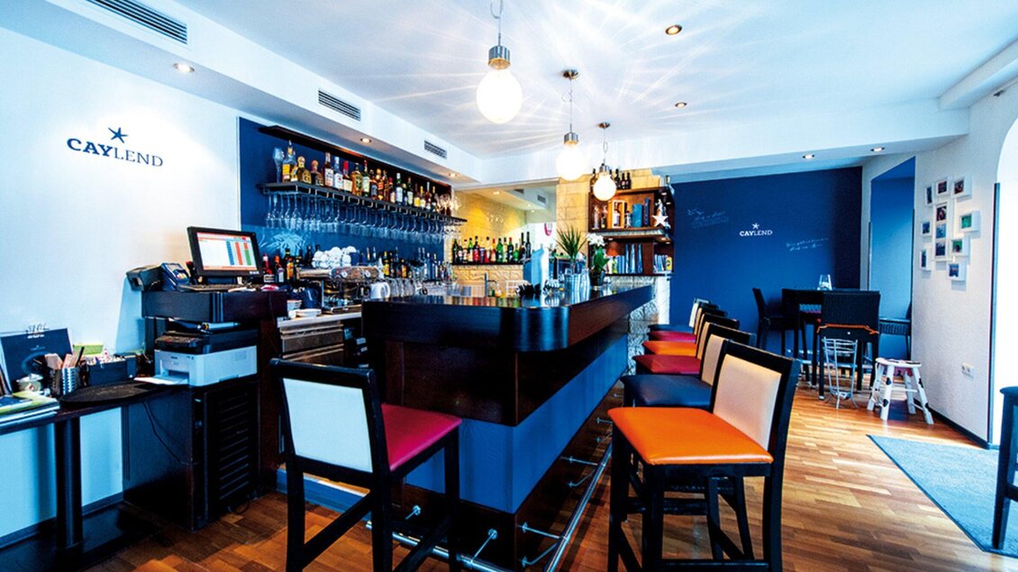 Eine stilvolle Bar mit einem eleganten Tresen und bunten Stühlen. Die Wände sind hell und die Beleuchtung sorgt für eine gemütliche Atmosphäre. | © Caylend - Michael Fiedler