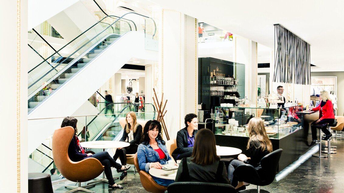 Ein eleganter Innenraum mit einer Cafébar und mehreren Sitzgruppen. Menschen entspannen sich und genießen ihre Zeit zwischen Treppen und stilvollem Dekor. | © Tageskaffee Freiblick