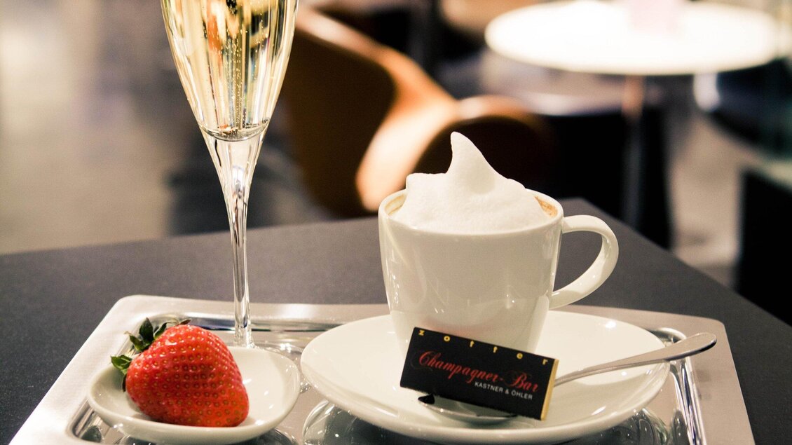Ein elegantes Tablett mit einem Glas Champagner, einer Tasse Kaffee mit Sahnehaube und einer Erdbeere. Der Hintergrund zeigt eine stilvolle Café-Atmosphäre. | © Tageskaffee Freiblick