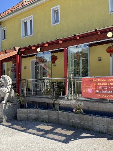 A Chinese restaurant with a large yellow facade and red lanterns. 

In front of the entrance, there are two lion statues and a banner for the grand reopening. | © TV Hochsteiermark