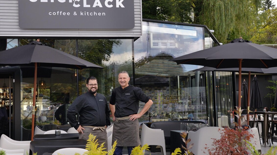 Zwei Männer stehen vor einem modernen Café mit dem Namen "CLACK coffee & kitchen". Der Außenbereich ist mit Tischen, Stühlen und Sonnenschirmen ausgestattet. | © Click Clack - Silvia Lambauer Fotografie